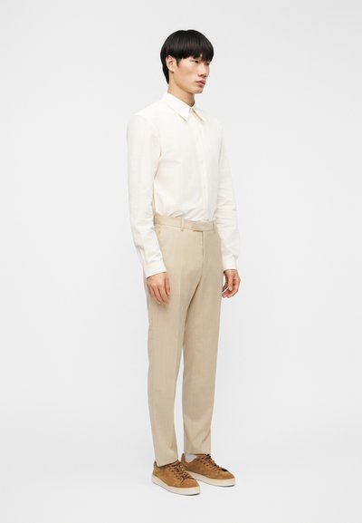 Φωτεινό μπεζ πουκάμισο με κουμπιά και μακρύ μανίκι, συνδυασμένο με πλισέ παντελόνια σε ανοιχτό μπεζ χρώμα και καφέ σουέντ αθλητικά παπούτσια. Απλός, εφαρμοστός σχεδιασμός με ομαλή υφή.