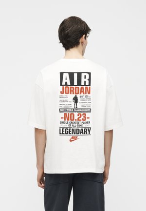 Άνδρας που φοράει λευκό φαρδύ T-shirt με μαύρο και πορτοκαλί κείμενο για την Air Jordan, συμπεριλαμβανομένου του "No. 23", επιτεύγματα και το λογότυπο της Nike στην πλάτη.