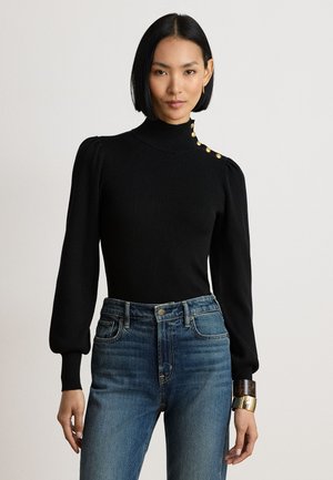 Femme aux cheveux noirs et raides porte un pull noir à col roulé à manches longues avec des boutons dorés et un jean bleu taille haute.