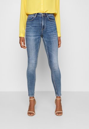 Femme portant un jean skinny bleu taille haute, un chemisier jaune à manches longues et des talons ouverts beiges, debout devant un fond blanc uni.