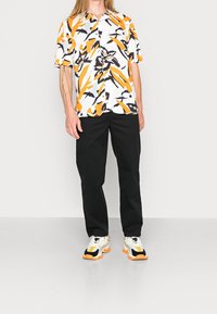 Short-sleeve, button-up shirt with an abstract orange, black, and white pattern. Paired with black trousers and multicolored sneakers.