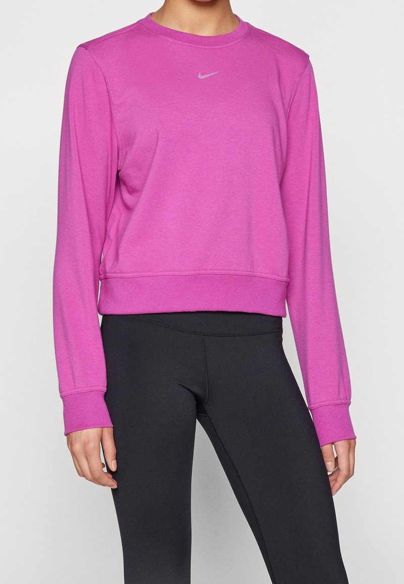 Personne portant un sweat-shirt Nike à manches longues rose vif et des leggings de sport noirs ajustés, debout devant un fond uni.