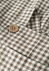 Brązowo-biała tkanina w kratkę gingham z drewnianym guzikiem, przedstawiająca prosty wzór, teksturę i detale szwów. Zbliżenie materiału odzieżowego.