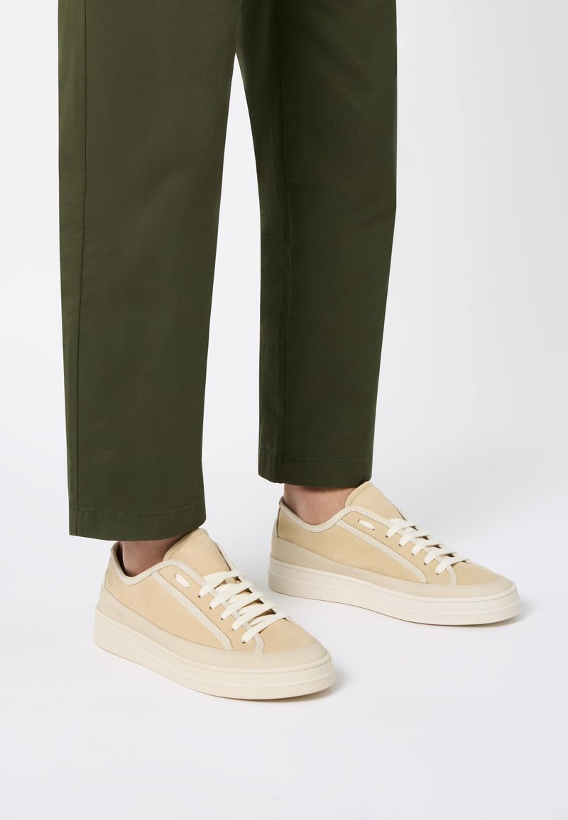Baskets beiges en matériaux suédés et en cuir, forme arrondie, semelle en caoutchouc blanche et fermeture à lacets. Associées à un pantalon vert à revers.