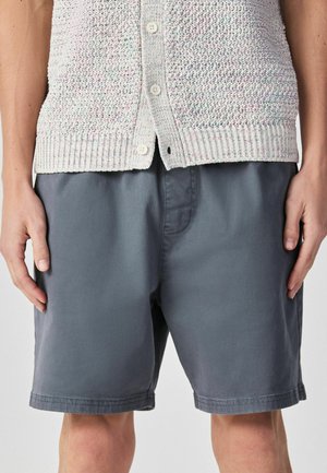 Közelkép egy személyről, aki világosszürke, texturált, gombos kardigánt és sötétszürke, térdig érő rövidnadrágot visel, kezei nyugodtan lelógatva oldalt.