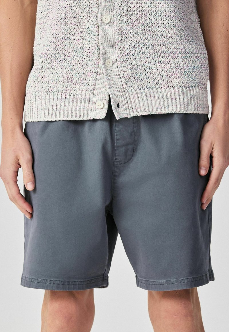 Gros plan sur une personne portant un cardigan boutonné texturé gris clair et un short gris foncé arrivant aux genoux, les mains détendues le long du corps.