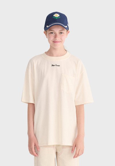 Ung person iført en cremefarvet oversized T-shirt med forlompe og en navy kasket med hvid blomst, smilende mod en ensfarvet baggrund.