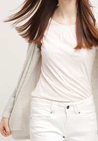 Blusa rosa chiaro con un sottile motivo floreale, abbinata a un cardigan crema e jeans bianchi. Tessuto liscio e dettagli con scollatura rotonda.