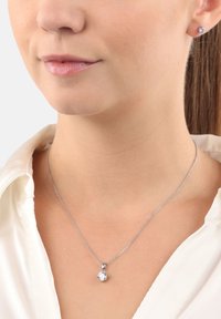 Silver necklace with a round pendant featuring a clear stone, worn with a white collared shirt. Simple chain design and minimal accents.