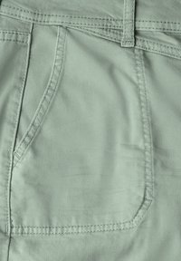 Tela de algodón verde claro que muestra un primer plano de un bolsillo trasero, con detalles de costura y una textura suave. El bolsillo tiene forma curva.