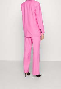 Completo tailored rosa composto da un lungo blazer con bottoni e pantaloni a gamba larga, realizzato in un tessuto liscio. Scarpe con tacco alto completano il look.