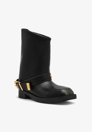 Bota de cuero negro para tobillo con tacón bajo, puntera redonda y correa decorativa con hebilla dorada alrededor del tobillo.