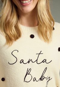 Kremasti pulover s crnim točkicama i izvezom "Santa Baby" u kurzivnom pismu. Mekana tekstura, okrugli izrez i dugi rukavi.