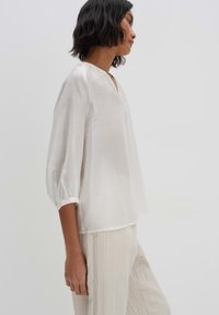 Blusa bianca a maniche lunghe con scollo a V, in tessuto leggero e maniche a sbuffo. Esposta su uno sfondo semplice per maggiore chiarezza.