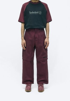 Man wearing black and burgundy Timberland T-shirt with matching burgundy cargo pants and boots, standing against white background.