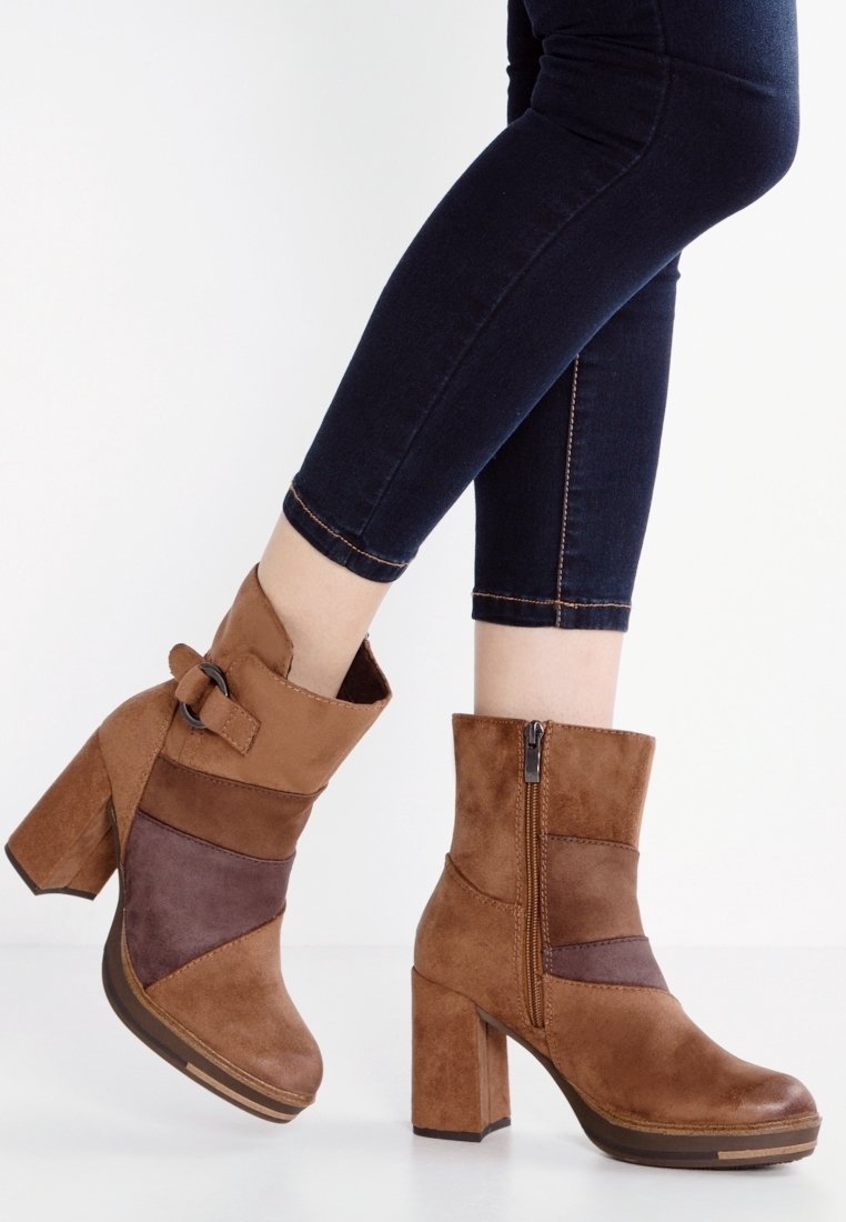 Jambes en jean skinny foncé portant des bottines en daim marron à talons carrés, fermeture éclair latérale et boucle décorative sur la sangle.