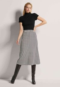 Schwarzes, geripptes Kurzarmoberteil kombiniert mit einem langen, schwarz-weißen Hahnentrittmuster-Rock; das Modell trägt kniehohe schwarze Stiefel.