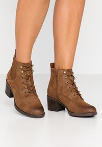 Bottes de cheville en suédine marron avec un laçage à l'avant, œillets dorés, fermetures éclair sur le côté et un talon empilé. Surface texturée et bout arrondi.