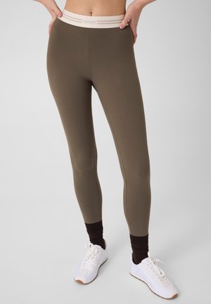 Leggings in olive green with beige elastic waistband labeled Guess, worn with black socks and white sneakers on a plain background.
