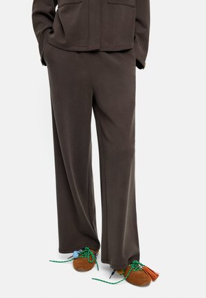 Osoba nosząca luźne ciemnobrązowe spodnie i brązowe sportowe buty z zielonymi sznurowadłami, ozdobione kolorowymi frędzlami i supełkami.