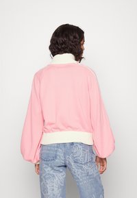 Rosa gerippter Pullover mit Ballonärmeln und weißem geripptem Kragen, mit einem taillierten Saum. Kombiniert mit hellblauen, gemusterten Jeans.