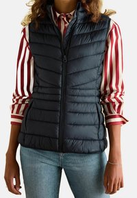 Granatowa kamizelka ocieplana z pikowanym wzorem, z przodem na zamek błyskawiczny i kieszeniami bocznymi, noszona na czerwono-białą w paski koszulę.