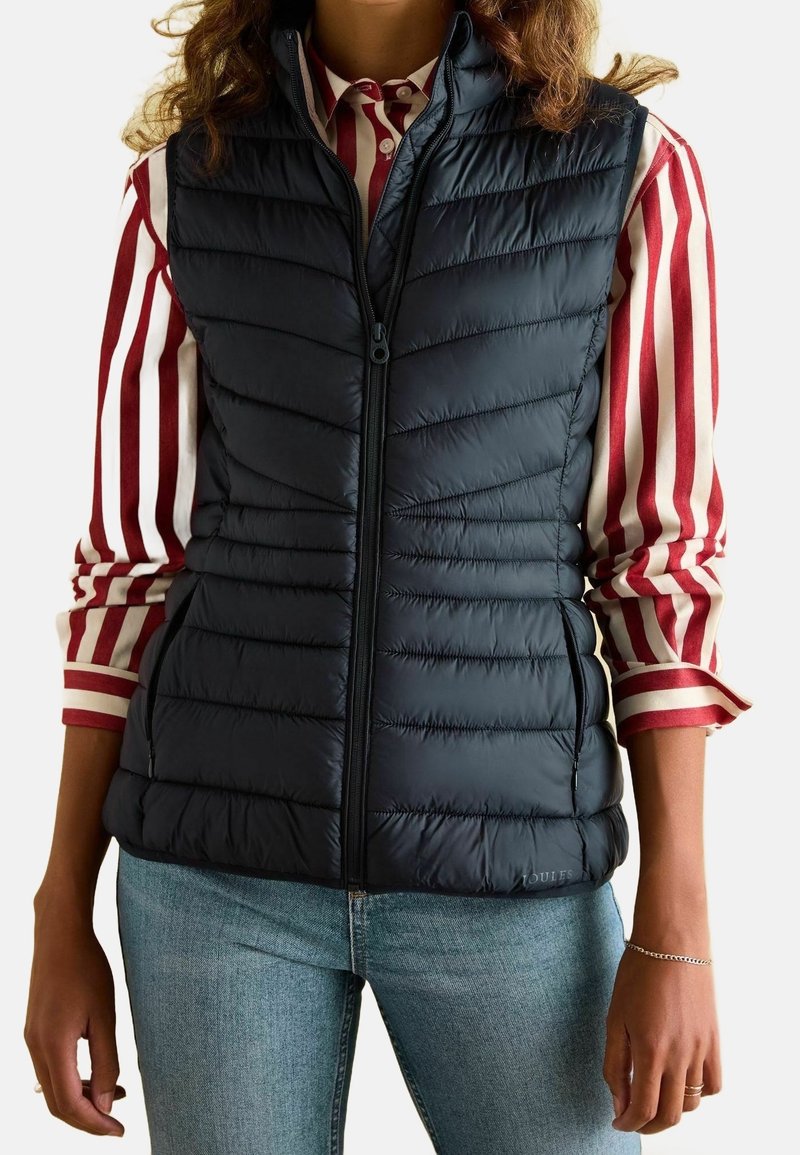 Granatowa kamizelka ocieplana z pikowanym wzorem, z przodem na zamek błyskawiczny i kieszeniami bocznymi, noszona na czerwono-białą w paski koszulę.