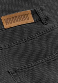 Gros plan d'une poche de pantalon noir texturé avec une étiquette en cuir marron de la marque Woodbird cousue près de la passants de ceinture.