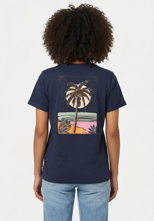 Persona con capelli ricci che indossa una maglietta blu navy con una grafica stilizzata di una palma e una spiaggia sul retro, abbinata a jeans azzurri.