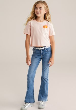 Lyserød cropped t-shirt med orange frugtgrafik, parret med lyseblå flared jeans og sølv sneakers, placeret mod en neutral baggrund.