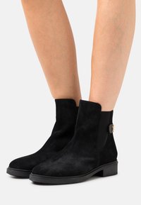 Botas de tobillo de ante negro con suela gruesa, paneles laterales elásticos y un pequeño detalle de hardware dorado cerca del tobillo para un uso fácil.