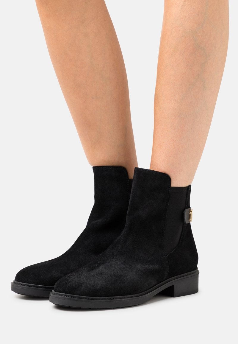 Botas de tobillo de ante negro con suela gruesa, paneles laterales elásticos y un pequeño detalle de hardware dorado cerca del tobillo para un uso fácil.