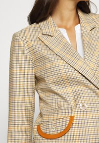 Kostkovaný blazer v béžové, hnědé a žluté barvě. Obsahuje klopové límce, dva knoflíky a zahnutý oranžový kapsový akcent. Texturovaná tkanina.