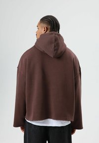 Sweat à capuche marron oversize avec une grande capuche, un ourlet brut et des manches longues, fabriqué en matériau doux. Porté par-dessus une chemise blanche et un pantalon foncé.