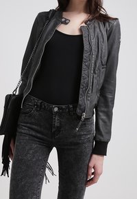 Donna che indossa una giacca di pelle nera sopra una canotta nera, abbinata a jeans neri a vita alta in lavaggio acido e che porta una borsa nera.