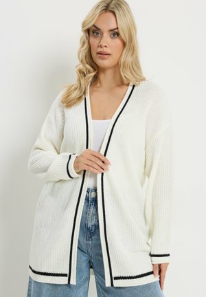 Blonde Frau trägt eine weiße Strickjacke mit schwarzem Besatz über einem weißen Oberteil und blauen Jeans, steht vor einem schlichten, hellen Hintergrund.