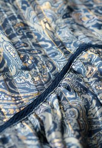 Tissu à motifs dans des tons bleus et dorés avec des dessins complexes. Présente des plis et une couture texturée le long de la couture centrale.