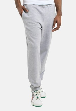 Man draagt lichtgrijze joggingbroek en witte sneakers met groene zolen, staat met één hand in de zak tegen een effen achtergrond.