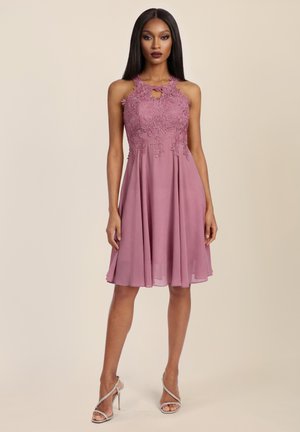 Frau in ärmellosem mauvefarbenem Kleid mit Spitzenoberteil und fließendem Rock, trägt silberne Riemchen-Sandalen mit hohem Absatz, steht vor einfachem Hintergrund.