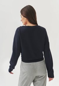 Námornícka mikina s dlhými, voľnými rukávmi a širokým výstrihom, s rebrovaným opaskom. Na sebe má svetlošedé elegantné nohavice.