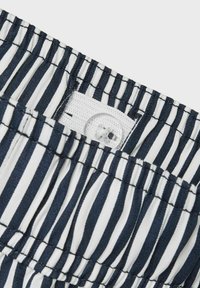 Tkanina w granatowo-białe paski z marszczoną strukturą i białym zapięciem na guzik. Materiał jest lekki i zaprojektowany z myślą o komforcie.