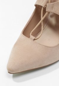 Chaussure plate beige à bout pointu en daim avec un fin détail de bride sur le cou-de-pied, présentant une texture lisse et un design minimaliste.