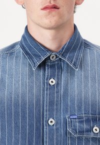 Blue denim shirt with white vertical stripes, buttoned placket, point collar, and a small pocket featuring a blue label. Textured fabric.