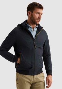 Mann trägt eine marineblaue Jacke mit Reißverschluss und gelben Details über einem hellblauen Hemd mit Knöpfen und beigen Hosen, steht mit einer Hand in der Tasche.