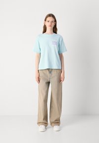 lichtblauw katoen t-shirt met korte mouwen en een grafisch logo. Gecombineerd met losse beige broeken en witte sneakers, staand tegen een neutrale achtergrond.