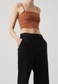 Zkrácený top s oranžovým a hnědým vlnovým vzorem, s nastavitelnými tenkými ramínky, v kombinaci s černými širokými kalhotami s kapsami.