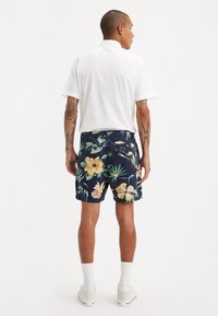 Cămașă albă cu mânecă scurtă combinată cu pantaloni scurți bleumarin cu modele florale mari în galben, verde și alb. Include șosete și pantofi albi.
