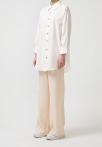 Personne portant une chemise blanche oversize à boutons, un pantalon crème large et des baskets blanches, debout devant un fond uni.