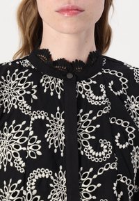 Blusa preta com bordados, apresentando motivos florais brancos, um gola alta de renda e fecho frontal de botões. Tecido suave com costura detalhada.