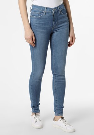 Personne portant un jean skinny bleu clair et des baskets blanches avec des détails à motifs, debout devant un fond clair uni.
